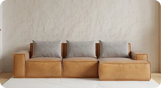 Immagine di un divano a tre posti sui toni del marrone e del grigio, poggiato su un tappeto bianco, con alle spalle un muro bianco dalla texture ruvida.
